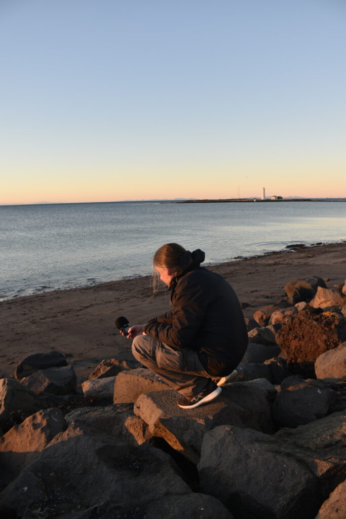 Balthazar Klimt crouching next to a lake, holding a recording device