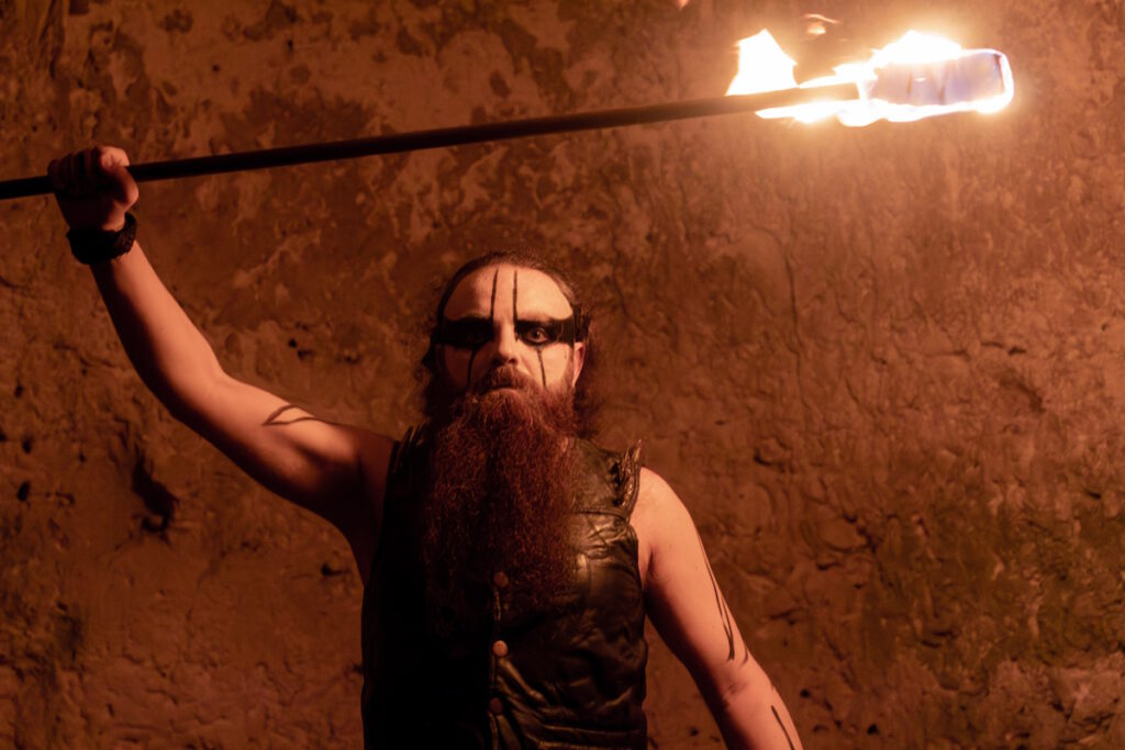Marton Saliba (Saħħar) standing in front of an ancient wall wearing corpsepaint, and holding a torch